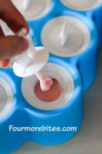 Stick for popsicle mold being placed into mold after strawberry liquid has been poured into a blue popsicle mold