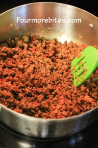 Cooked ground beef, ground pork sausage, onions, peppers and spices in a stainless steel pot on a black stovetop