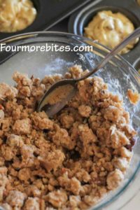 Streusel topping mixed in a clear glass bowl with a spoon in the bowl.  There are muffin tins in the background that have already been filled with muffin batter.