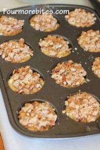 Dark muffin pan on a white counter top.  Each muffin cup is filled with batter and then streusel is sprinkled on top in each muffin cup.